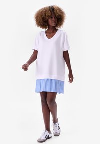 Losse witte top met een V-hals en opgerolde mouwen, gecombineerd met een lichtblauwe geplooide rok met verticale strepen.