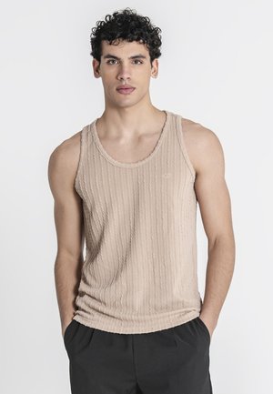 Ung mand med krøllet mørkt hår iført en beige tekstureret tanktop og sorte bukser, stående med hænderne i lommerne mod en hvid baggrund.