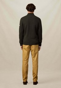 Tmavě zelený pletený svetr s vysokým límcem, žebrovanými rukávy a lemem, kombinovaný s béžovými kalhotami a hnědými botami, pohled zezadu.