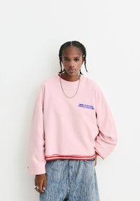 Felpa rosa dal taglio rilassato, con testo blu sul petto. Abbinata a jeans a gamba larga blu chiaro e una collana in argento.