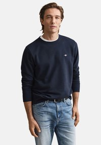 Marineblå sweatshirt med rund halsudskæring, ribstrikkede manchetter og et lille logo på brystet, kombineret med lyseblå jeans og et sort bælte.