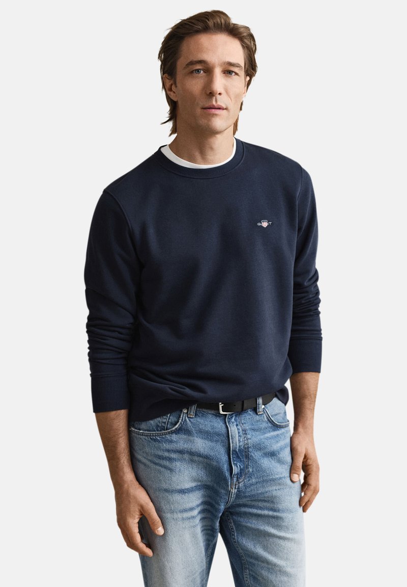 Marineblå sweatshirt med rund halsudskæring, ribstrikkede manchetter og et lille logo på brystet, kombineret med lyseblå jeans og et sort bælte.