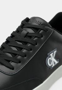 Κοντινή λήψη μαύρου δερμάτινου αθλητικού παπουτσιού με μαύρα κορδόνια, γκρι και λευκό λογότυπο "cK" στο πλάι και λεπτομέρεια με το λογότυπο στη γλώσσα.
