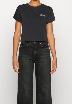 Osoba ubrana w czarną, skróconą koszulkę Levi's oraz czarne, wysoko osadzone szerokie spodnie na tle jednolitego białego tła.