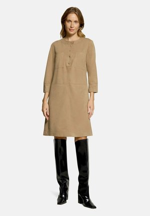 Kvinde iført en beige langærmet knælængde kjole med knapper ved brystet og sorte knæhøje hæle støvler, der står mod en ensfarvet baggrund.