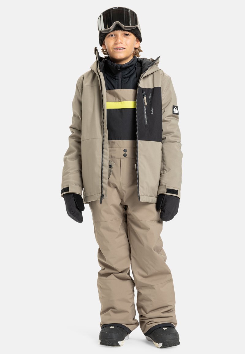 Ensemble de veste et pantalon de ski, beige avec des accents noirs et jaunes. Fermeture éclair, plusieurs poches et gants noirs. Chaud et isolé.