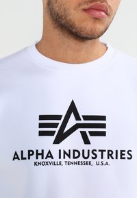 Witte sweatshirt met een zwart gedrukt logo met lijnen en een "A" ontwerp, samen met de tekst "ALPHA INDUSTRIES, KNOXVILLE, TENNESSEE, U.S.A."
