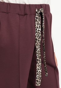 Pantalons bourgogne avec une taille froncée et un cordon de serrage à imprimé léopard. Texture lisse et détail de couture latérale visibles.