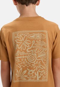 Camiseta de algodón marrón con un gráfico inspirado en la naturaleza de color crema en la parte posterior, que representa plantas y un diseño en espiral.