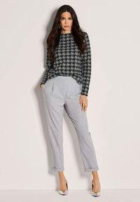Vrouw met lang donker haar, gekleed in een zwart-grijze houndstooth trui, lichtgrijze omgeslagen broek en zilveren hoge hakken, staande met handen in de zakken.