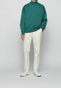 Pull col roulé vert en tricot, associé à un pantalon sur mesure crème et des chaussures blanches à semelle épaisse.