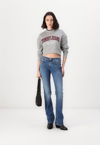 Felpa grigia corta con "TOMMY JEANS New York" stampato in rosso, abbinata a jeans a campana blu e a una piccola borsa a tracolla nera.