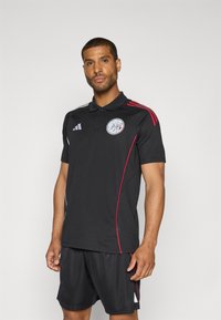 Sort polo shirt med et rundt logo og røde detaljer, parret med sorte shorts. Materialet ser let og åndbart ud.