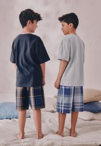 Des garçons portent un t-shirt bleu marine avec un short à carreaux et un t-shirt gris avec un short à carreaux bleu, debout dos à dos sur un lit avec des coussins.