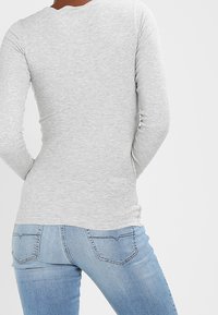 Maglietta grigia chiara a maniche lunghe con design aderente, tessuto a coste texturato, scollatura rotonda e orlo posteriore leggermente curvato, abbinata a jeans blu.
