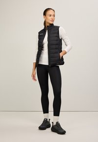 Schwarze gesteppte Weste mit hohem Kragen, getragen über einem weißen Langarmshirt, kombiniert mit schwarzen Leggings und dunklen Schuhen. Verfügt über Seitentaschen.