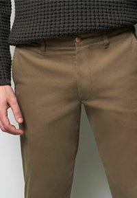Bruine katoenbroek met een gladde textuur, ritsgulp, knoopsluiting en zijzakken. De hand rust gedeeltelijk zichtbaar op de dij.