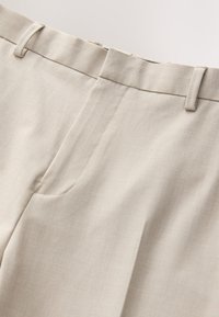 Gros plan d’un pantalon en tissu beige montrant la ceinture, les passants et la braguette avant avec un tissage texturé subtil.