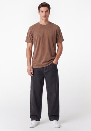 Man staat recht voor zich uit kijkend, draagt een bruin T-shirt met korte mouwen, losse zwarte broek, witte sneakers, met één hand in zijn zak en een neutrale gezichtsuitdrukking.