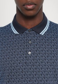 Μπλούζα polo σε ναυτικό μπλε χρώμα με επαναλαμβανόμενο ανοιχτό μπλε μοτίβο 'MK', διαθέτει γιακά και δύο κουμπιά. Ραβδωτές πινελιές στον γιακά.