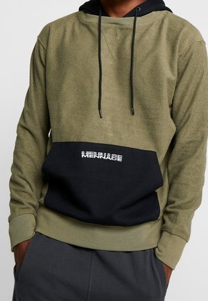 Muž v olivově zelené texturované mikině s kapucí, s černou přední kapsou a logem "MENNACE", ruce v kapsách tmavě šedých kalhot.