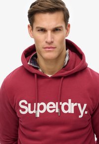 Maroon hoodie with a large cracked white "Superdry." logo. Features a drawstring hood and a soft fabric texture.