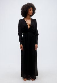 Γυναίκα με μαλλιά σε σγουρά, φορώντας μακρύ μαύρο φόρεμα με βαθύ V στο ντεκολτέ και φουσκωτά μακριά μανίκια, στέκεται ξυπόλυτη μπροστά σε απλό φόντο.