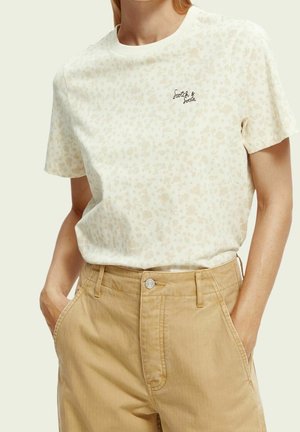 Nainen käyttää vaalean beigeä, kukkakuviollista T-paitaa, jossa lukee "Scotch & Soda", ja vaaleanruskeita, korkeavyötäröisiä housuja, joiden taskuissa on kädet.