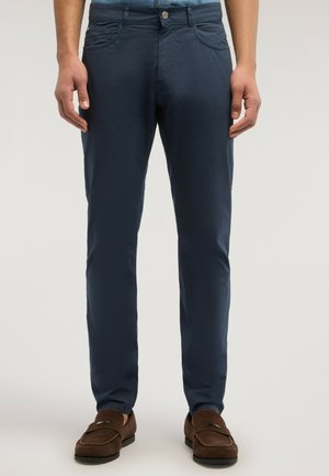 Mand iført mørkeblå slim-fit bukser og brune ruskindsløbere, stående mod en ensartet lys grå baggrund.