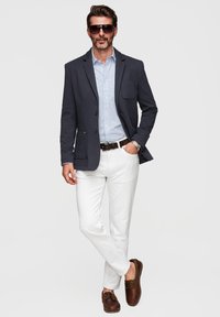 Homme portant un blazer sombre, une chemise bleue rayée, un pantalon blanc, une ceinture marron, des mocassins marron, une montre et des lunettes de soleil, debout avec une main dans la poche.
