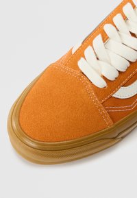 Baskets en suède orange avec lacets blancs, bande latérale blanche et semelle en caoutchouc translucide couleur gomme, gros plan sur la zone des orteils.