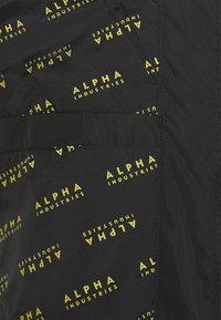 Tissu noir avec le texte "ALPHA INDUSTRIES" répété en jaune vif. Présente une texture lisse et une poche latérale pour plus de praticité.