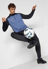 Sporty sæt med en blå og sort langærmet trøje med lynlås, sorte leggings og sorte sko, sammen med en mønstret fodbold.