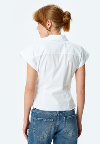 Camicia bianca a maniche corte con colletto, vestibilità aderente e texture liscia; abbinata a jeans blu con vita alta e tasche posteriori.