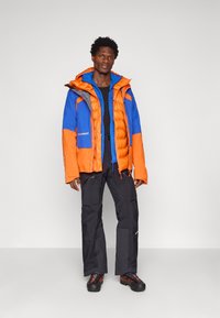 Niebieska i pomarańczowa puchowa kurtka z kapturem, czarne spodnie i teksturowane obuwie. Kurtka ma zamek z przodu i detal z logo.