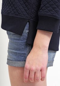 Navy quilted sweatshirt with ribbed cuffs, paired with light blue denim shorts. Visible hand resting on the shorts, showcasing detail.