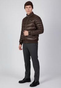 Chaqueta acolchada marrón con cremallera, cuello alto y parche con logo, combinada con pantalones grises y zapatos negros; textura suave, diseño acolchado.