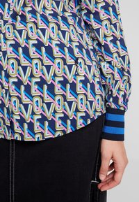 Monivärinen nappipaita, jossa toistuva "LOVE"-grafiikka keltaisena, pinkkinä, sinisenä ja vihreänä tummansinisen pohjan päällä; raidalliset hihansuut.