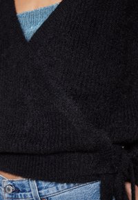 Gros plan d’un pull en tricot noir à nouer sur le côté, porté sur un top bleu et associé à un jean en denim bleu.
