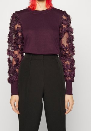 Personne portant un haut violet foncé avec des manches transparentes à volants et un pantalon noir taille haute, debout devant un fond uni.