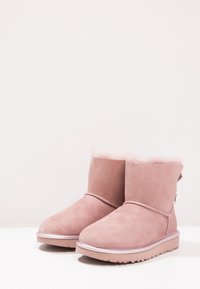 Różowe zamszowe botki za kostkę z puszystą wyściółką wewnętrzną, teksturowanym zewnętrzem i płaską gumową podeszwą. Posiadają ozdobny tylny uchwyt.