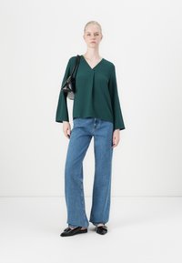 Kvinna i mörkgrön V-ringad blus, vida blå jeans, svarta spetsiga loafers och svart axelväska som står mot en vit bakgrund.