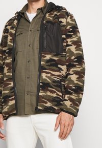 Fleecová bunda s maskáčovým vzorem, černou nylonovou náprsní kapsou a zipem vpředu. Má kapuci a žebrované manžety, volný střih.