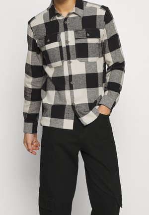 Mann, der ein schwarz-beiges kariertes Hemd mit langen Ärmeln, zwei Brusttaschen und schwarze Cargohosen trägt.