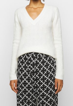 Femme portant un pull blanc texturé à col en V avec des manches longues et un pantalon noir avec un motif géométrique blanc.