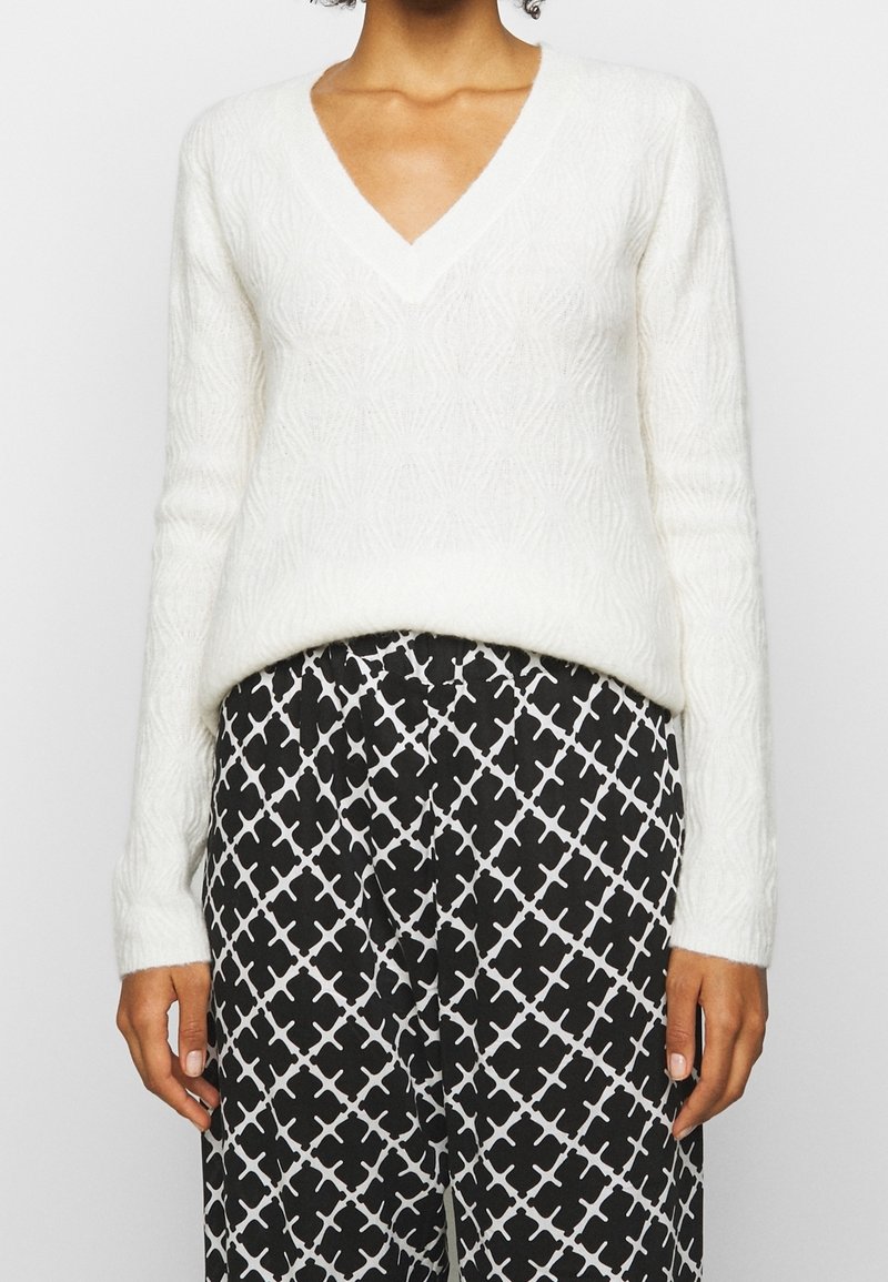 Femme portant un pull blanc texturé à col en V avec des manches longues et un pantalon noir avec un motif géométrique blanc.