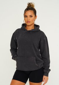 Dunkelgrauer, übergroßer Hoodie aus weichem Stoff mit einer Fronttasche, Kapuze mit Zugband und gerippten Bündchen. Kombiniert mit schwarzen Shorts.