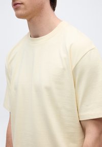 Άνδρας που φοράει απλό, κοντομάνικο, ανοιχτό κίτρινο T-shirt, εμφανιζόμενος από τον λαιμό έως το μέσο του κορμού του, μπροστά σε απλό φόντο.