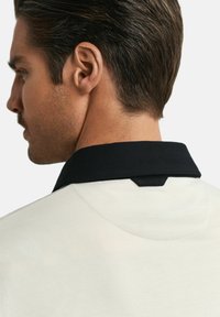 Polo pour homme avec un corps crème et un col noir, finition texturée et une étiquette de marque à l'arrière du col.