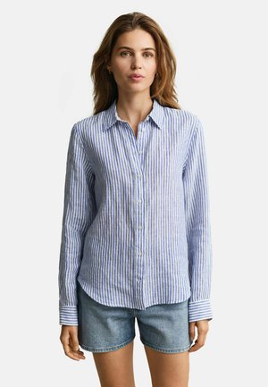 Vrouw met een blauw-wit gestreept overhemd met lange mouwen en blauwe spijkershorts, staand tegen een effen witte achtergrond.